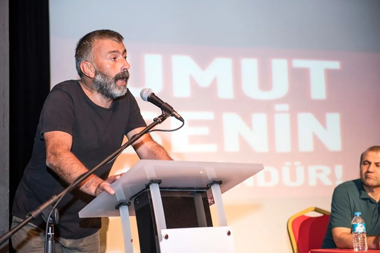 Başaran Aksu speaking at a podium with a microphone, a Turkish-language Umut-Sen union banner projected on the wall behind him