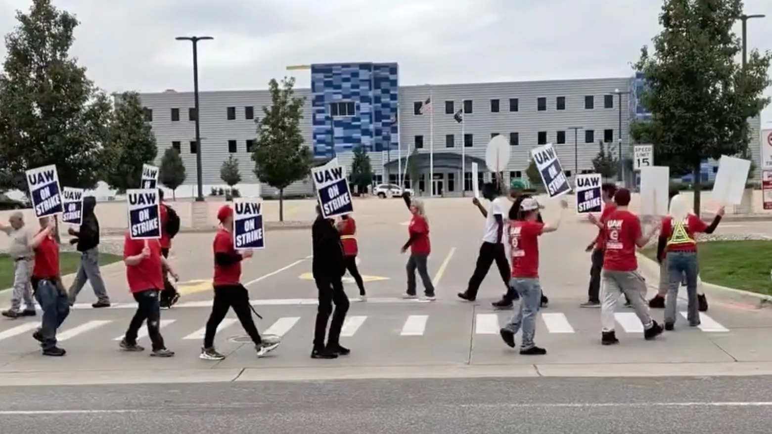 UAW workers in red picket shirts carrying “UAW ON STRIKE” signs, marching across a crosswalk in front of an office building.