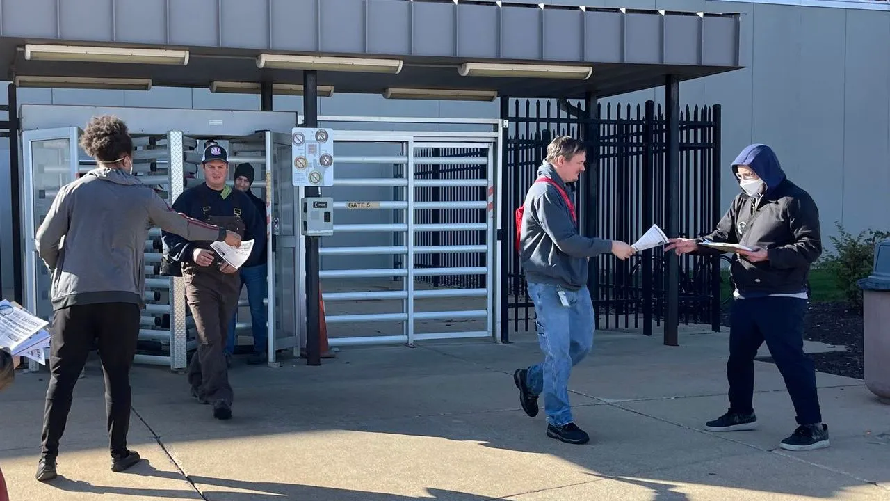 Campaign supporters distribute Will Lehman for UAW President leaflets to autoworkers at a plant entrance marked Gate 5, beside metal security turnstiles and black perimeter fencing on a sunny day.