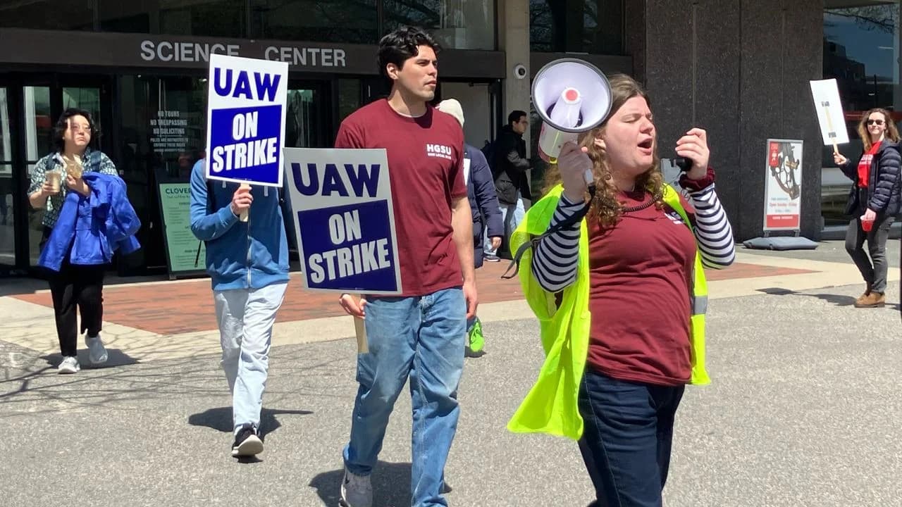 Harvard graduate student workers picketing in front of the Harvard Science Center, holding blue “UAW ON STRIKE” signs, with one worker in a yellow safety vest leading chants through a megaphone