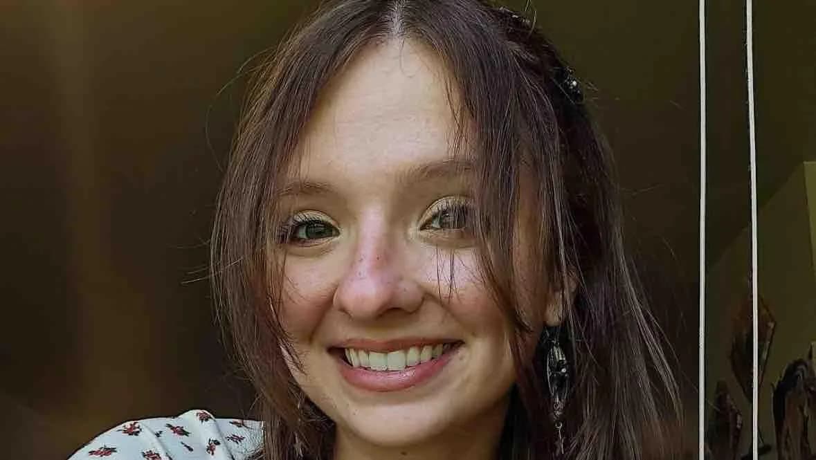 Chloe Freeman in a close-up photo, smiling, with shoulder-length brown hair, a dark hair clip and dangling earrings, wearing a white top with a small red floral print.