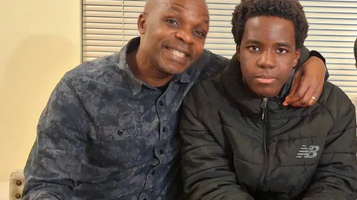 Kennedy Orwa, a man in a blue patterned shirt, with his arm around his teenage son who is wearing a black New Balance jacket