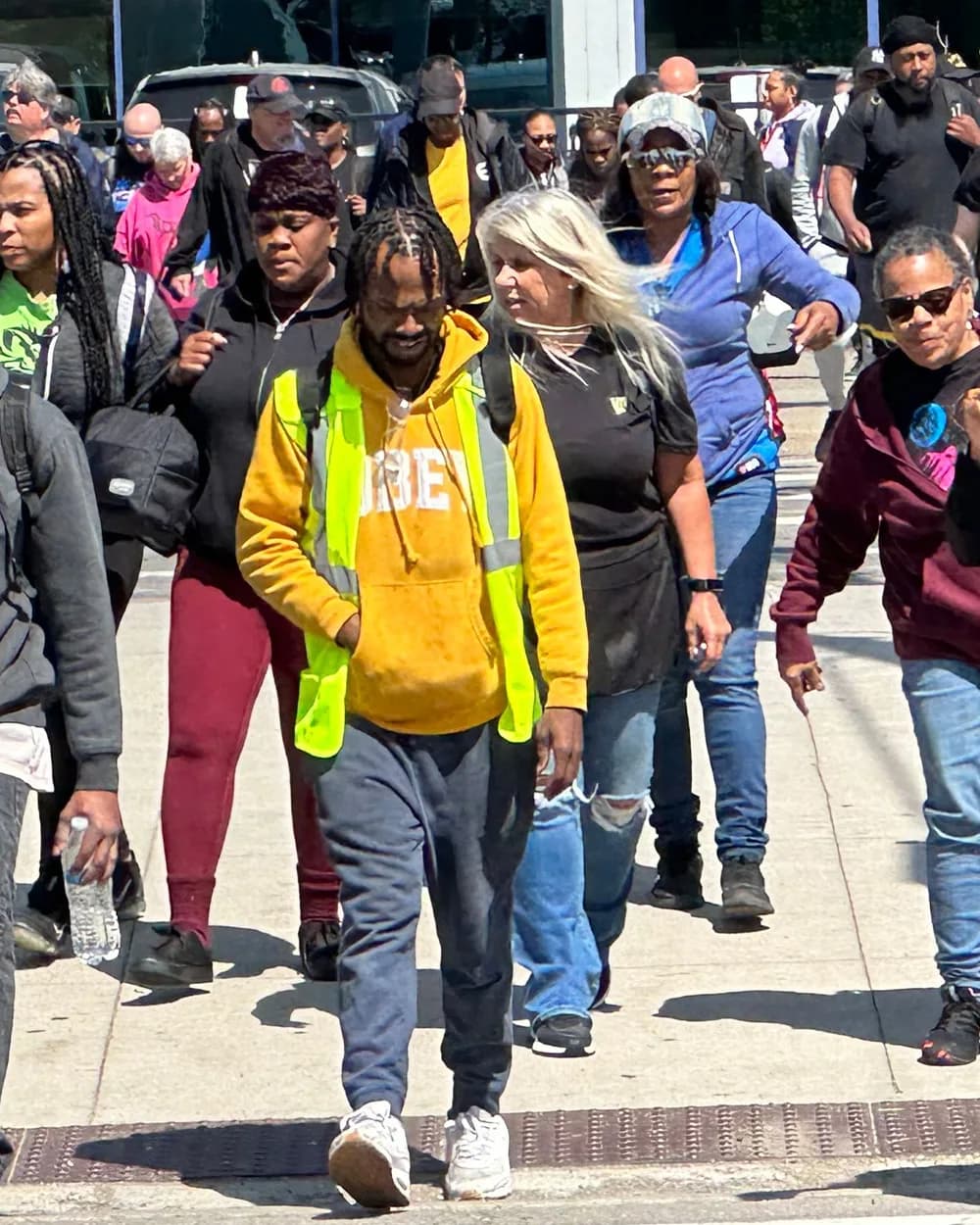 A crowd of workers walking together on a city street, led by a man in a yellow safety vest and orange hoodie.