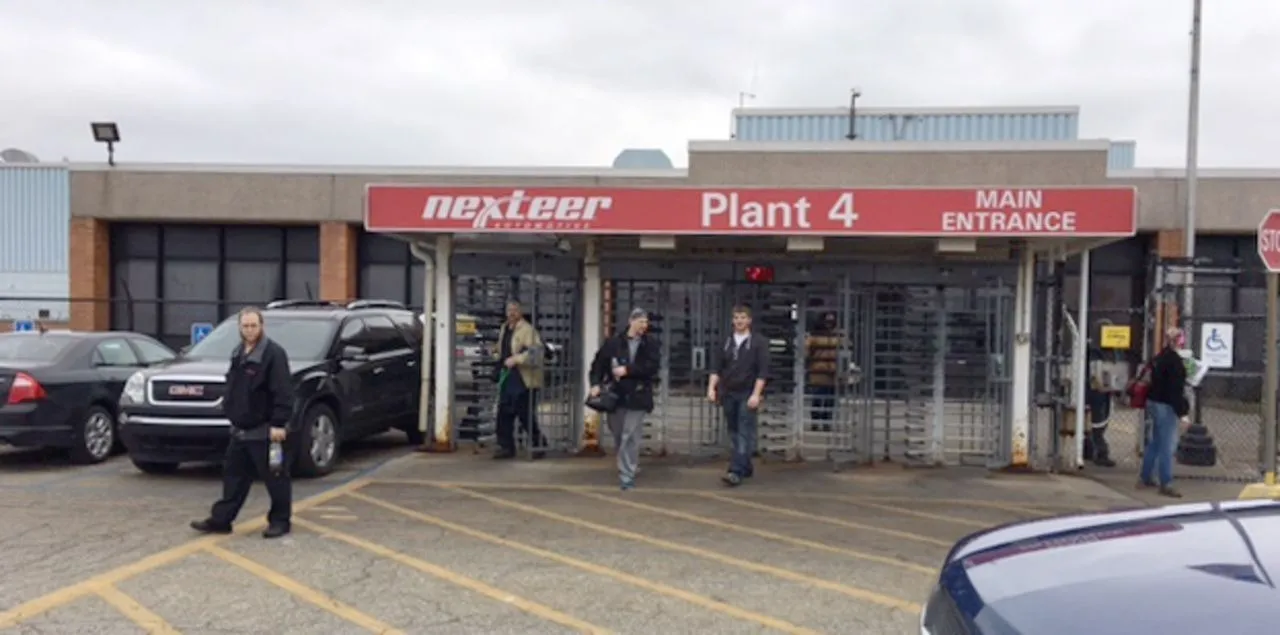Workers walking out through turnstiles at the Nexteer Automotive Plant 4 Main Entrance in Saginaw, Michigan, with a red Nexteer sign overhead reading Plant 4 Main Entrance