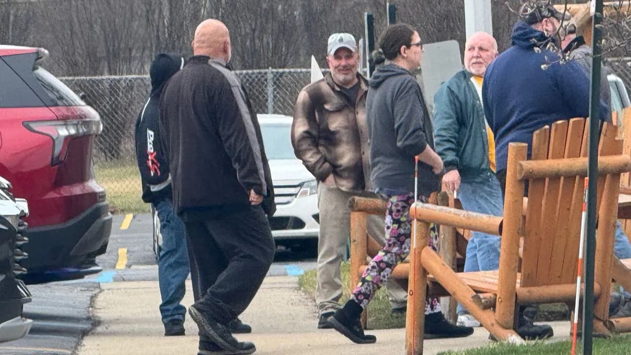 A group of Nexteer auto parts workers in casual jackets gathered outdoors in a parking lot beside a chain-link fence, with cars and wooden log chairs nearby, one worker in a brown jacket and white cap smiling toward the camera