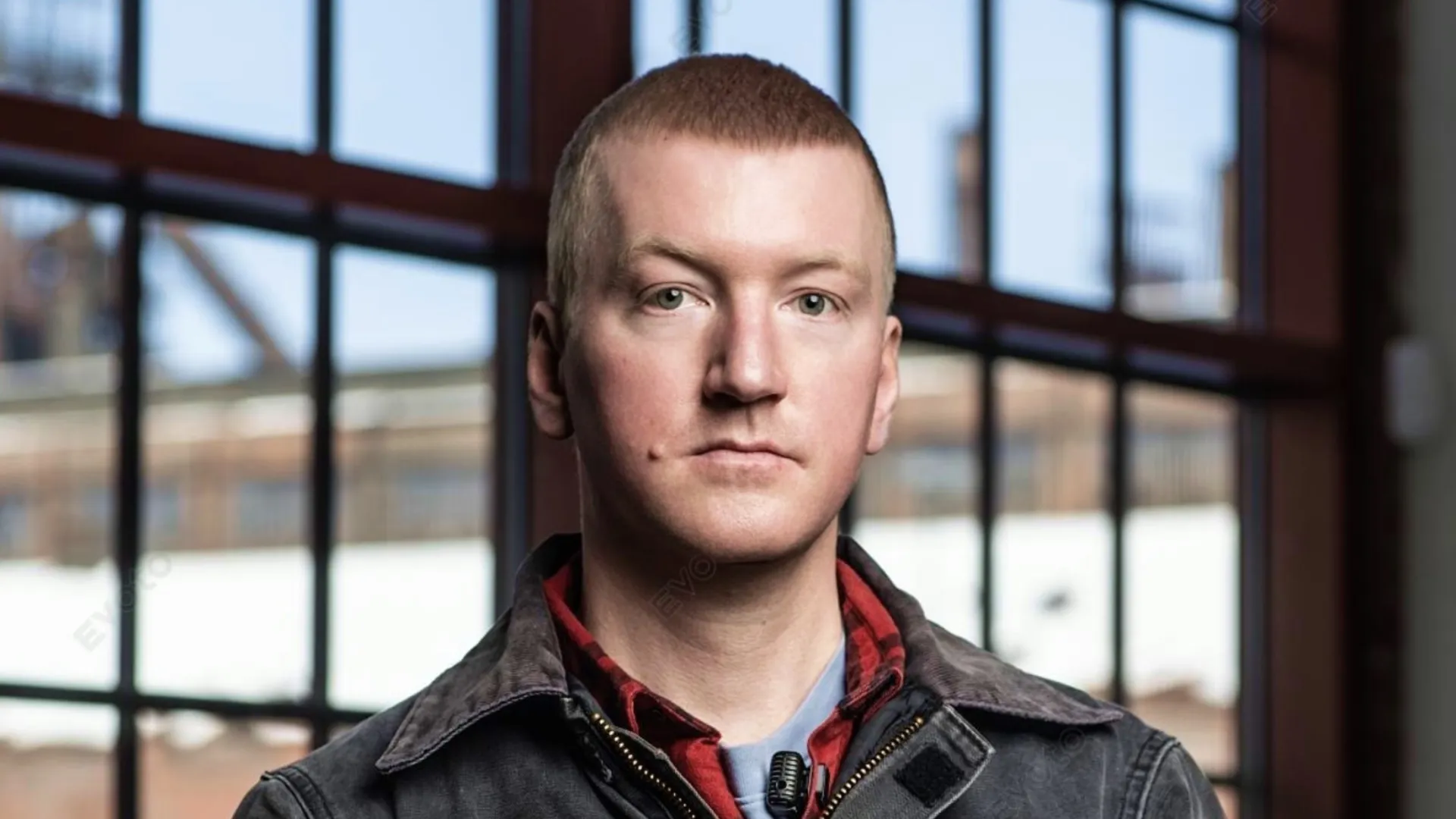 Will Lehman, a young man with short light hair in a gray jacket and red flannel shirt, standing in front of large industrial windows with an industrial building visible in the background
