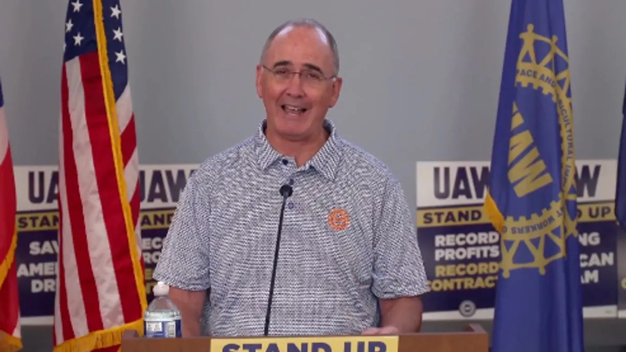 Shawn Fain speaking at a podium flanked by American and UAW flags, with “STAND UP” campaign signage behind him