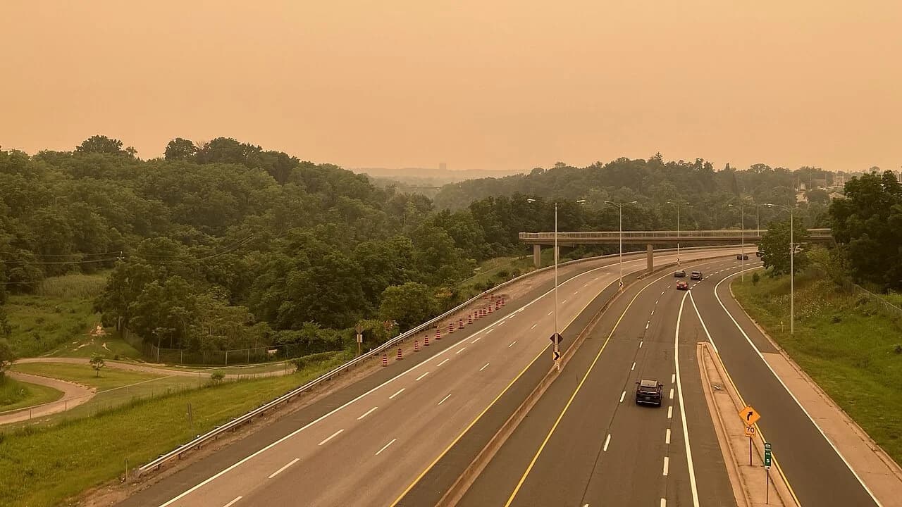 A multi-lane highway shrouded in thick, orange-tinted wildfire smoke haze, with a distant overpass, scattered cars, and construction cones along the shoulder; the sky and tree line are obscured by smog.