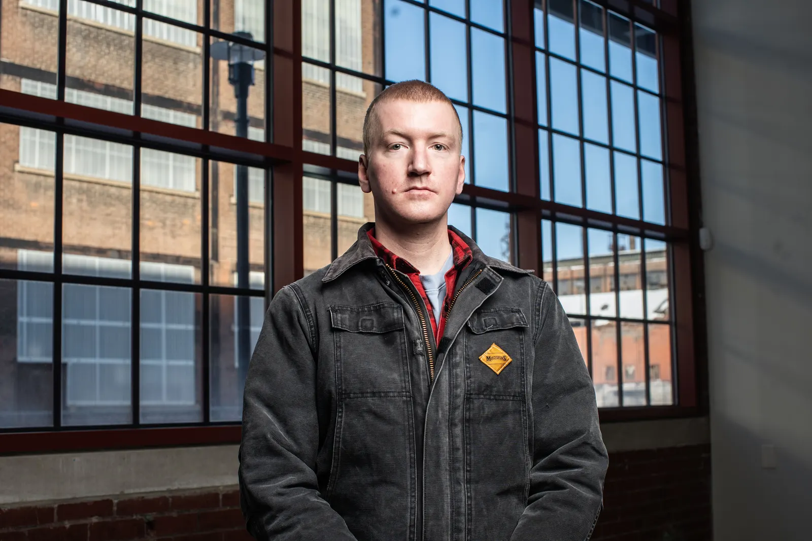 Will Lehman standing in a dark work jacket over a red flannel shirt, with a yellow Muleskins patch on the chest, in front of tall factory windows that look out on brick industrial buildings.