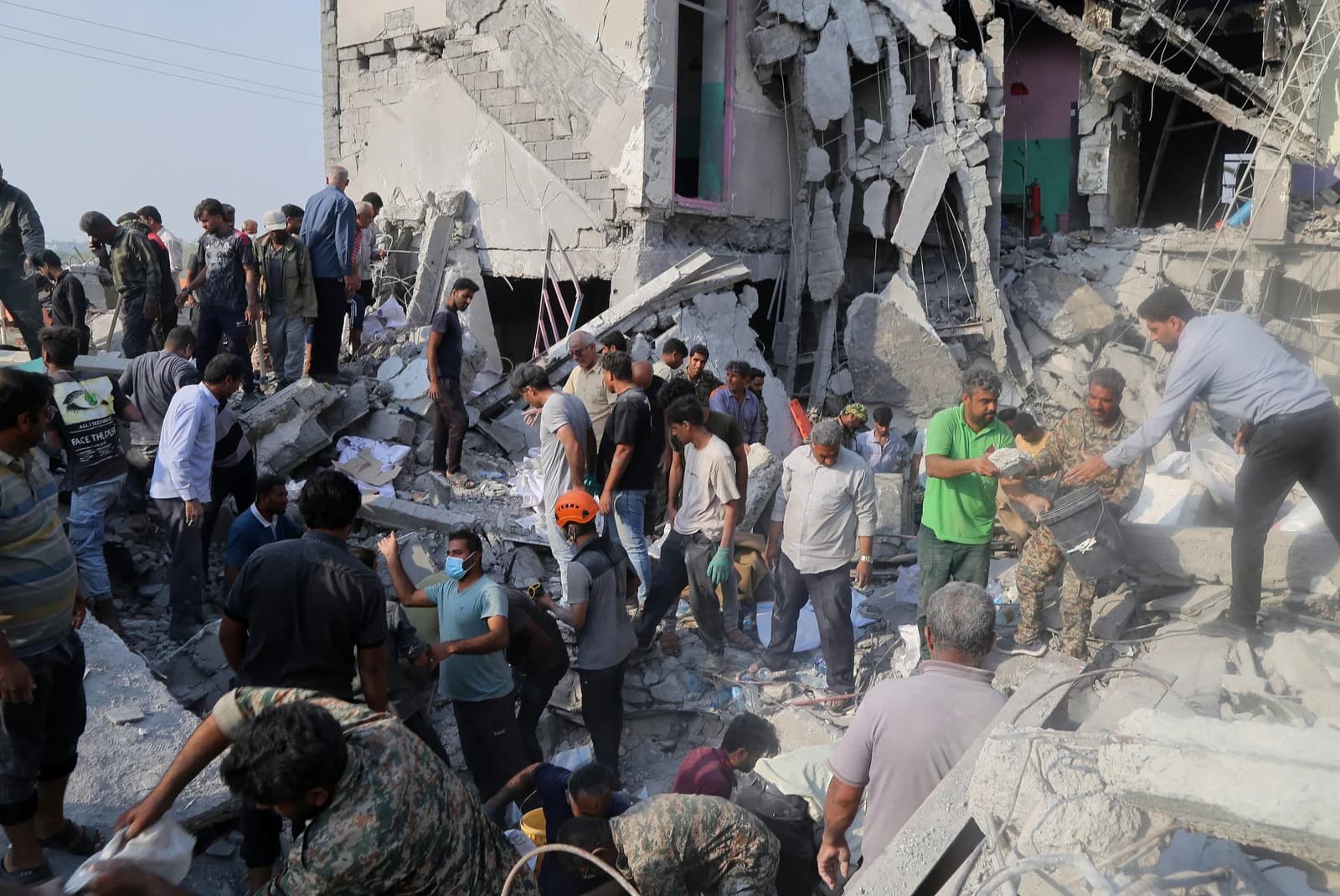 Civilians and rescue workers in red helmets searching through the rubble of the bombed Shajareh Tayyebeh school in Minab, Iran
