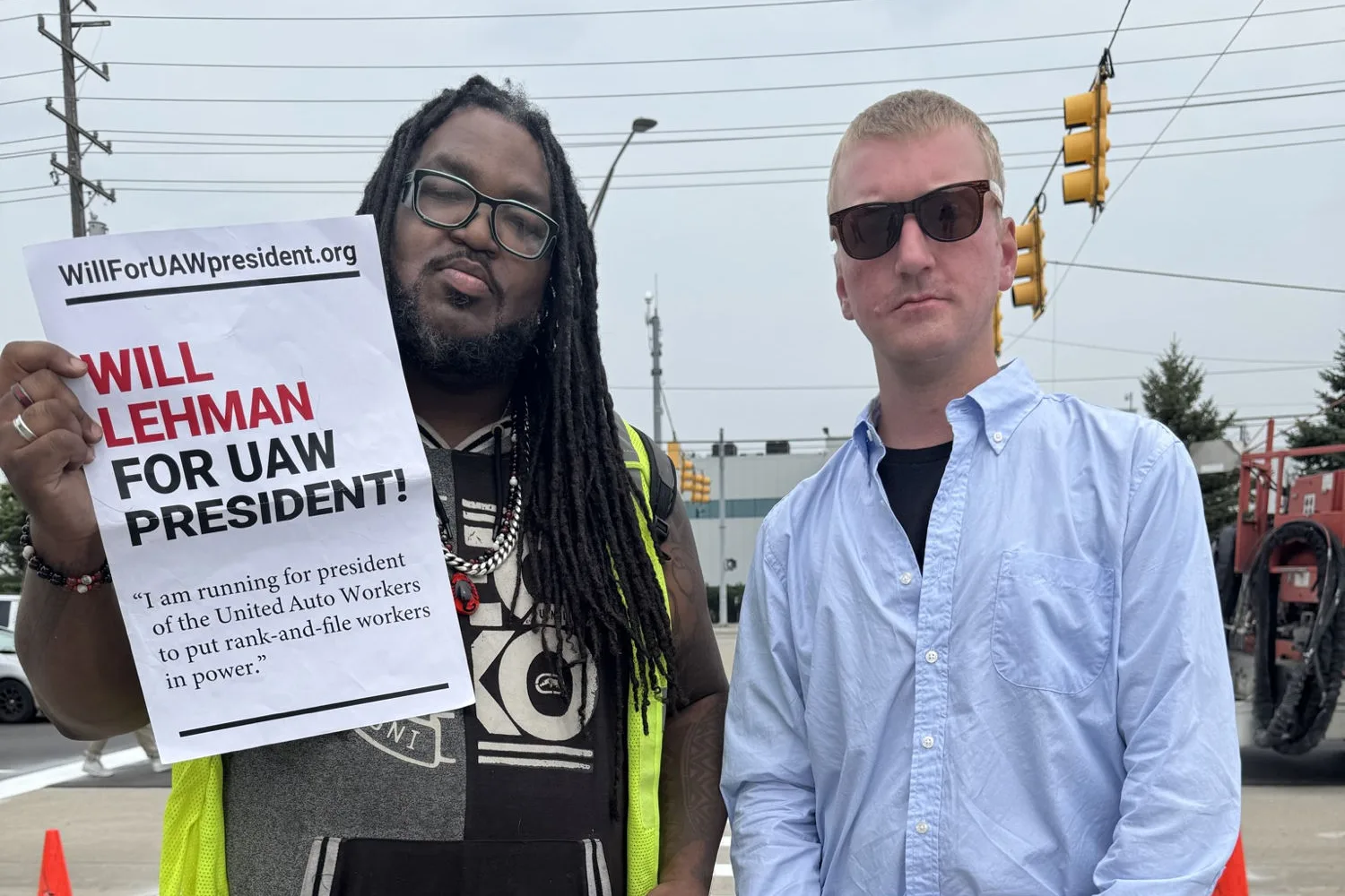 A worker in a hi-vis vest holds up a “WILL LEHMAN FOR UAW PRESIDENT!” poster while standing next to Will Lehman at an entrance to the Stellantis Warren Truck Assembly Plant, with traffic signals and a factory building behind them.